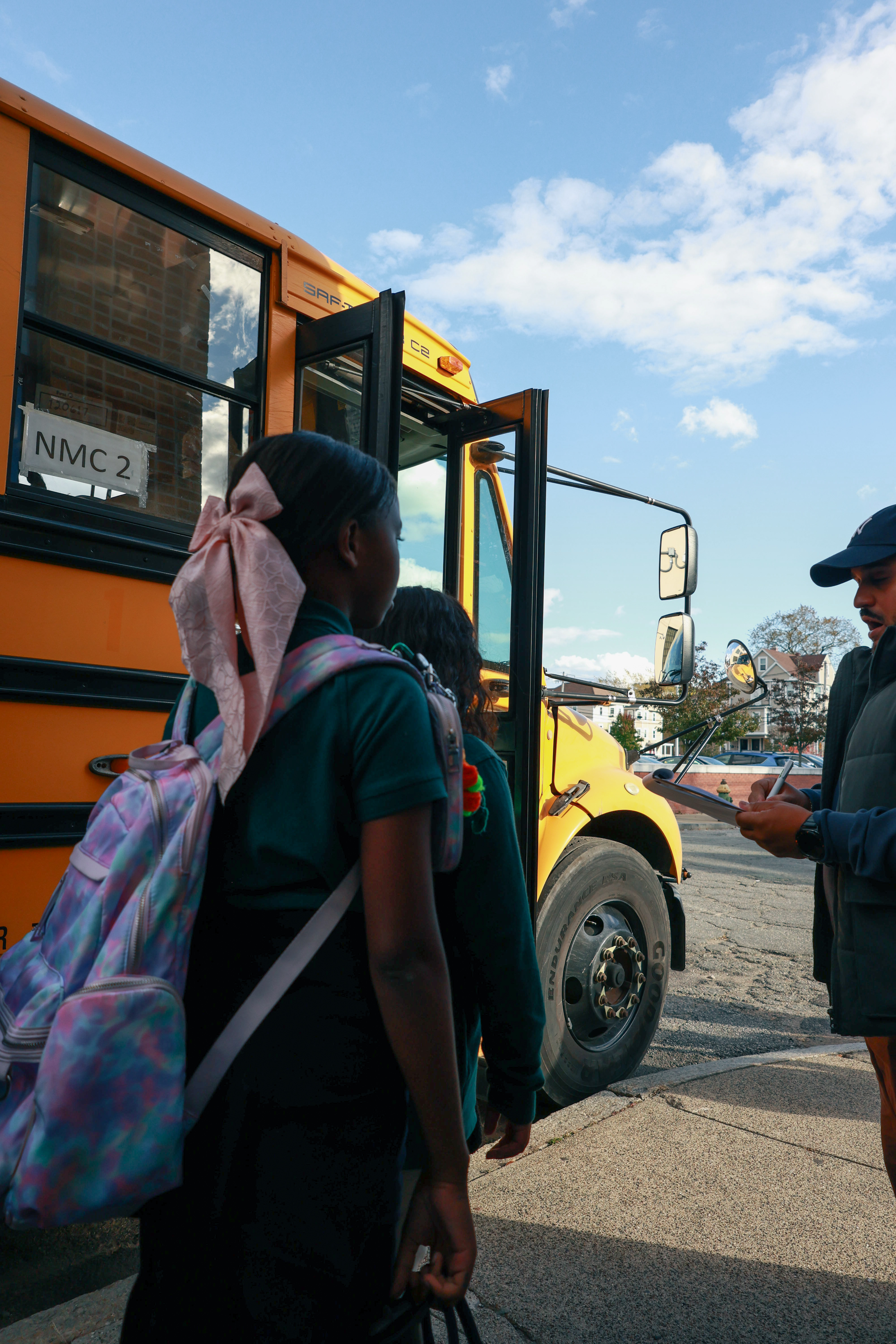 A girl with a light pink bow and tie-dye back pack stands waiting to board a yellow school bus.